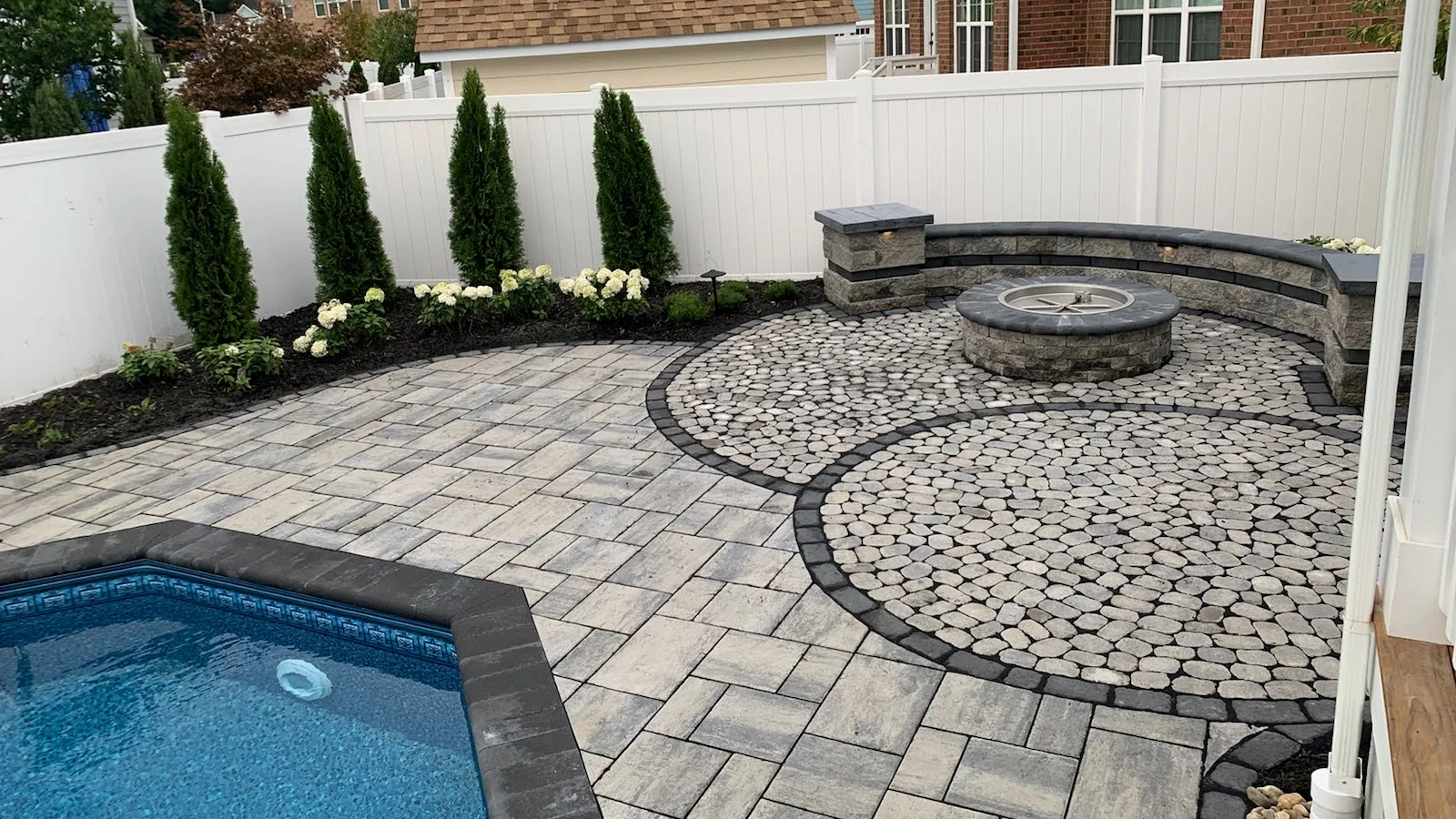 Finished stage: completed paver terrace with a pool, fire pit, seat wall, and planting