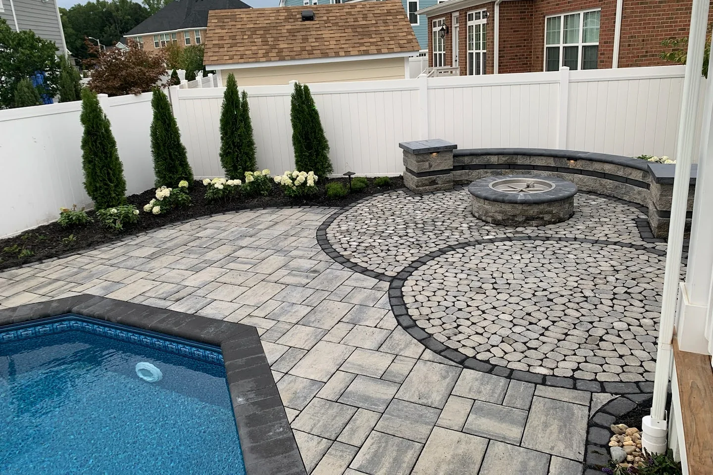 Completed residential paver patio with a pool, fire pit, seat wall, and planting beds